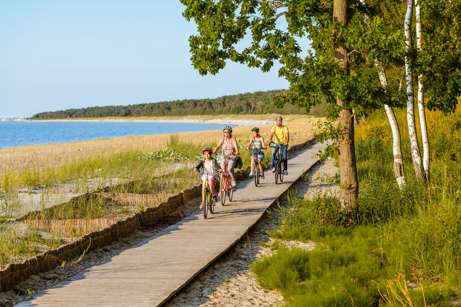 Familie fährt mit Fahrrädern über Holzweg am Lubminer Strand. Links ist der Strand und das Wasser, rechts stehen Bäume.